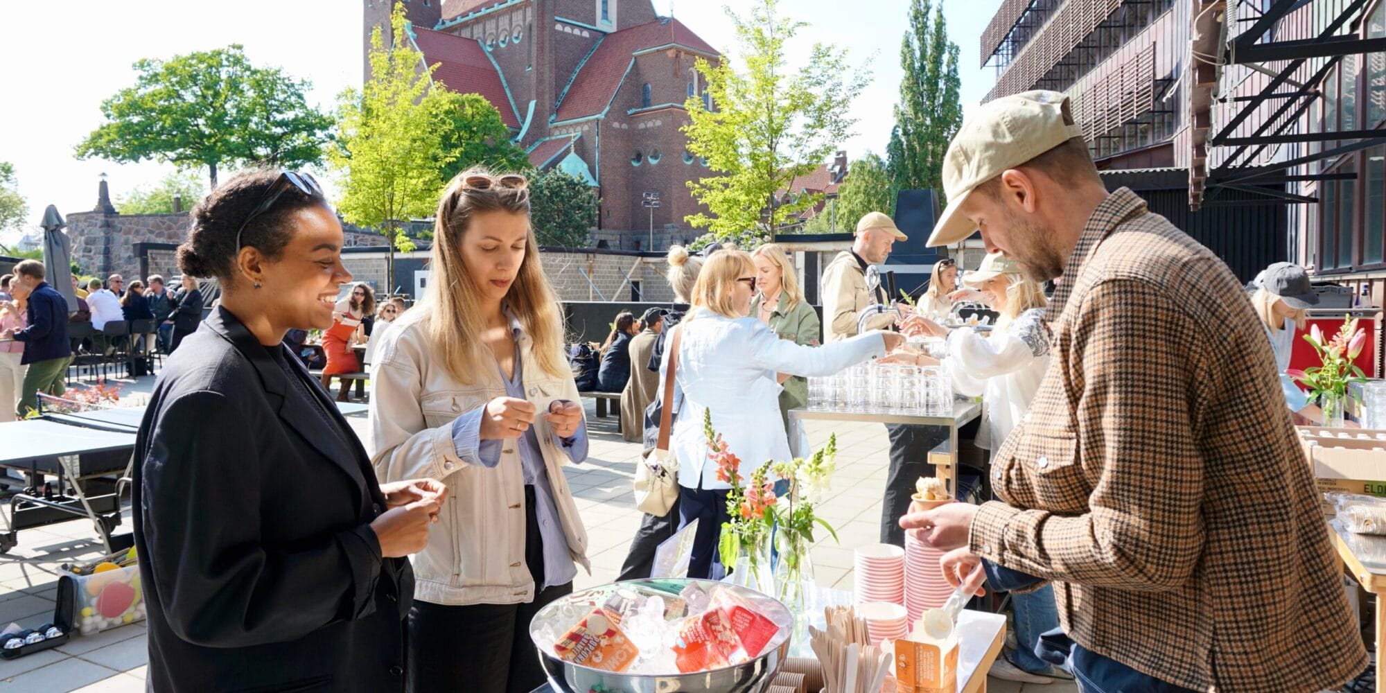 Utomhusevenemang med människor vid en servering, kyrka i bakgrunden och grönskande träd i soligt väder.