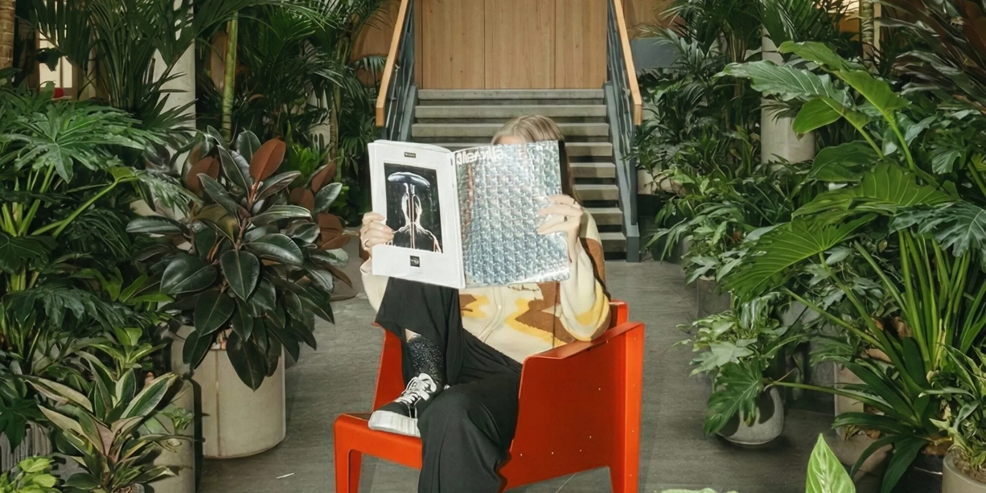 En stor entré beklädd i växtlighet och grönska. I förgrunden sitter en person i en röd plastfåtölj och håller en tidning framför ansiktet. 